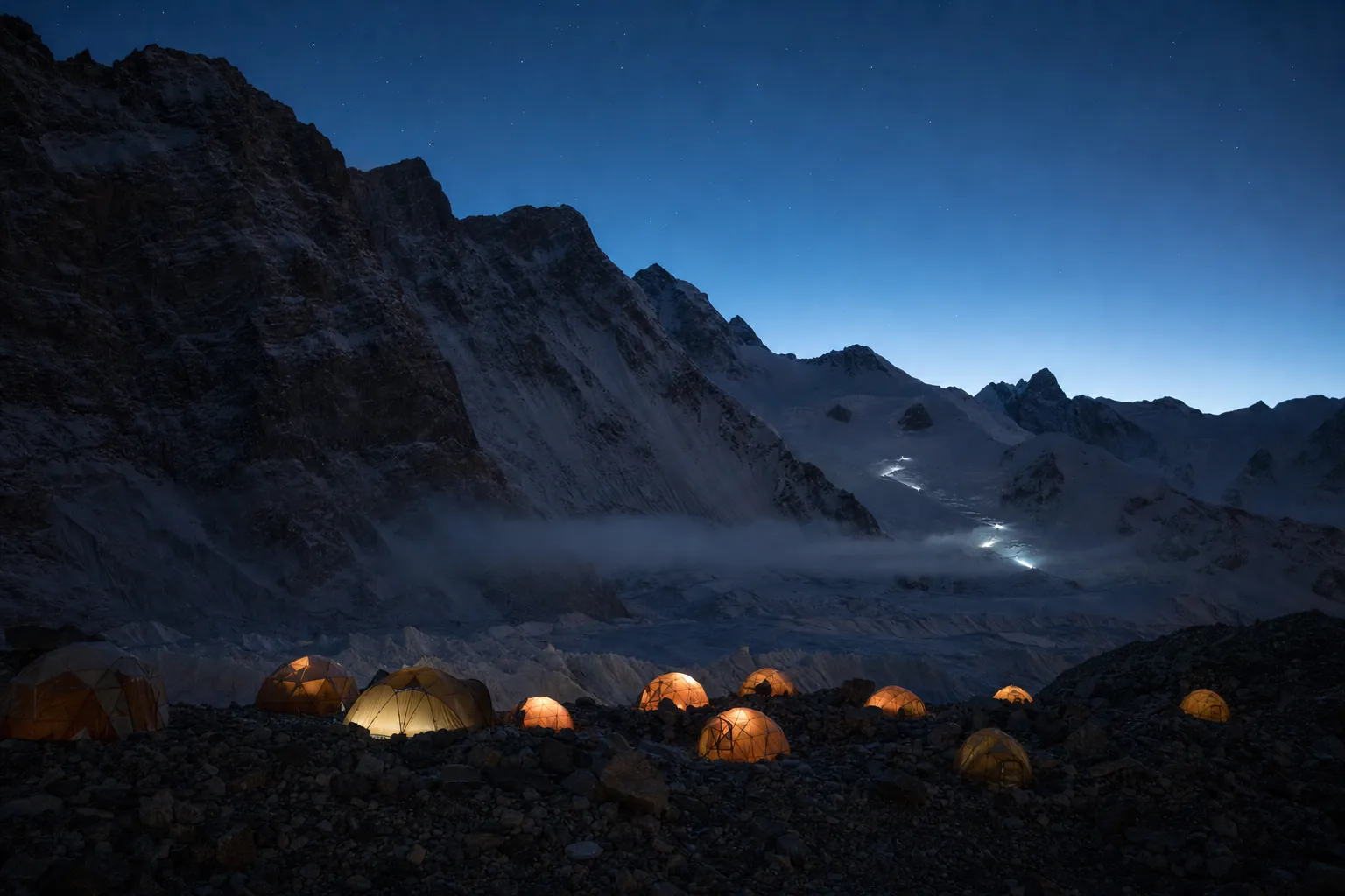 Expedition tents glowing at blue hour beneath a dark mountain wall, headlamps on the glacier